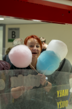 Uśmiechnięta słuchaczka o rudych włosach trzyma w dłoniach trzy duże waty cukrowe w kolorach różowym, niebieskim i białym, patrząc w stronę obiektywu zza szyby.
