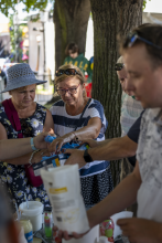 Grupa seniorów na świeżym powietrzu skupiona przy stoisku warsztatowym, z zaciekawieniem oglądająca produkty i materiały podawane przez prowadzącego.